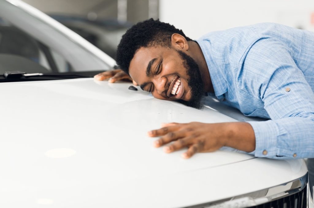 New Car Owner. Happy African American Man Touching Hugging His Brand-New Auto Cherfully Buying Vehicle In Auto Dealership. Selective Focus